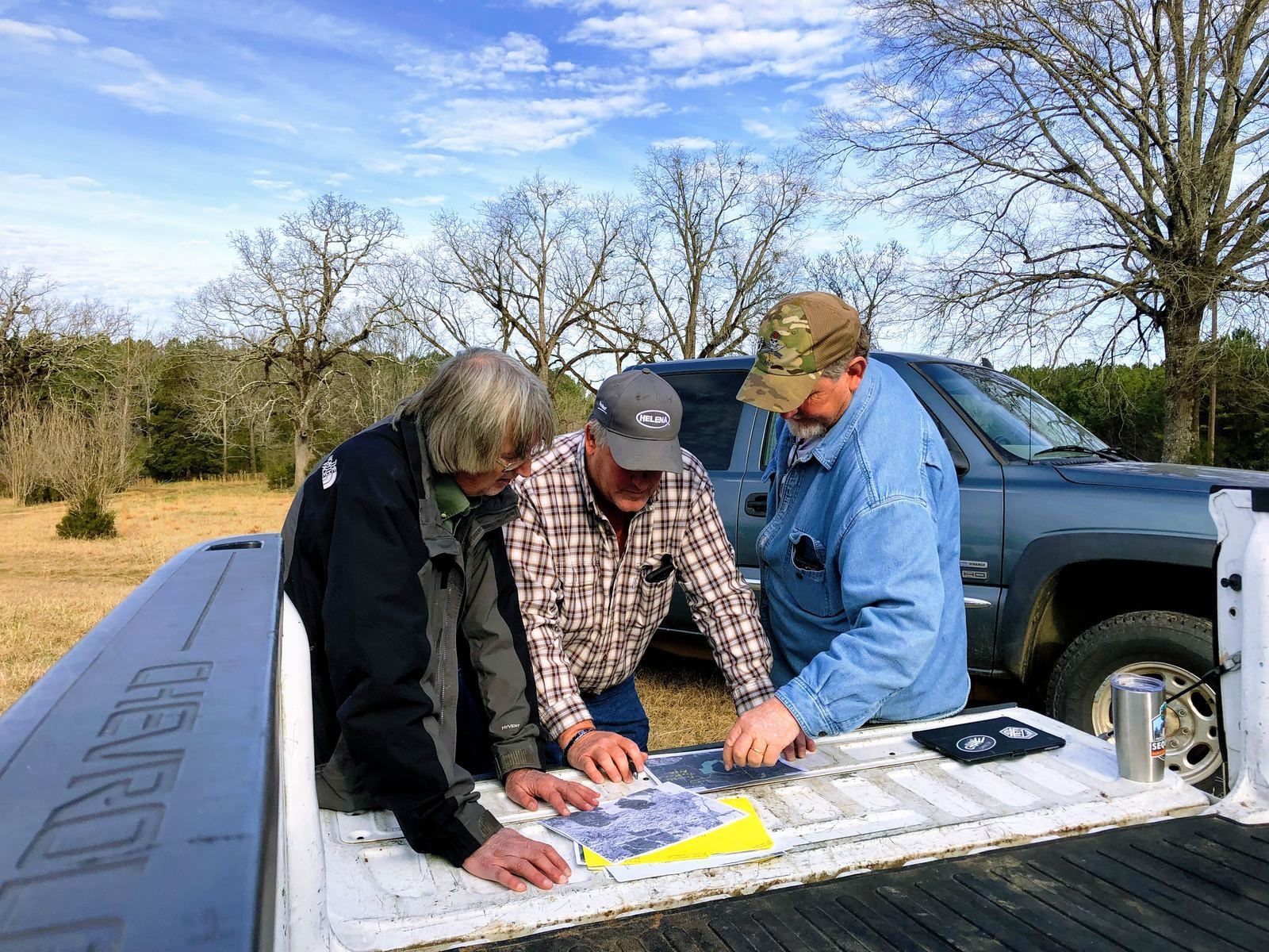 Three men going over a tract map spread on a pickup tailgate at the edge of a field